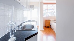 Una cocina moderna con armarios blancos, encimera negra y un lavabo. Se ve una ventana con persianas en el fondo, y una cama con una mesita de noche de madera en la habitación contigua.