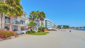 Un hotel frente a la playa con edificios de varios pisos, palmeras y un paseo arenoso que conduce a la orilla.