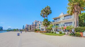 Un hotel frente a la playa con varios balcones, palmeras y un paseo marítimo de arena.