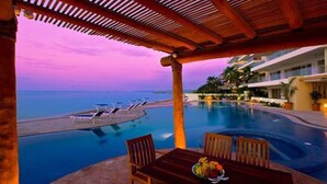 A poolside area with a view of the ocean at dusk, a dining table with chairs, and a fruit bowl. - Best value !! Luxury Beach Front Condo (Emiliano Zapata)