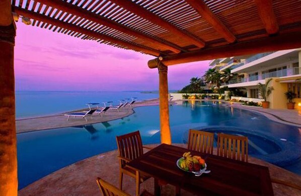 A poolside area with a view of the ocean at dusk, a dining table with chairs, and a fruit bowl.