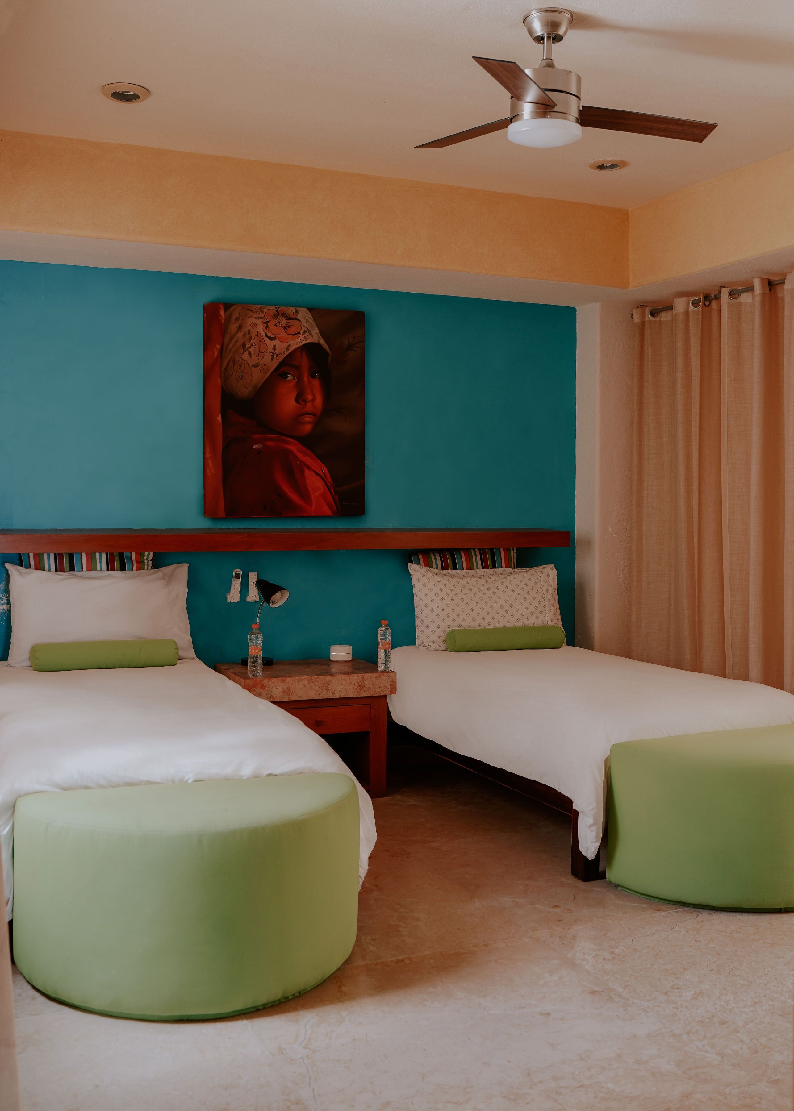 A hotel room with two beds, a ceiling fan, a painting on the wall, and a window with curtains.