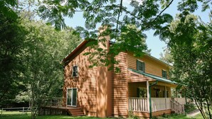 A wooden cabin with a porch, surrounded by trees and a grassy yard.