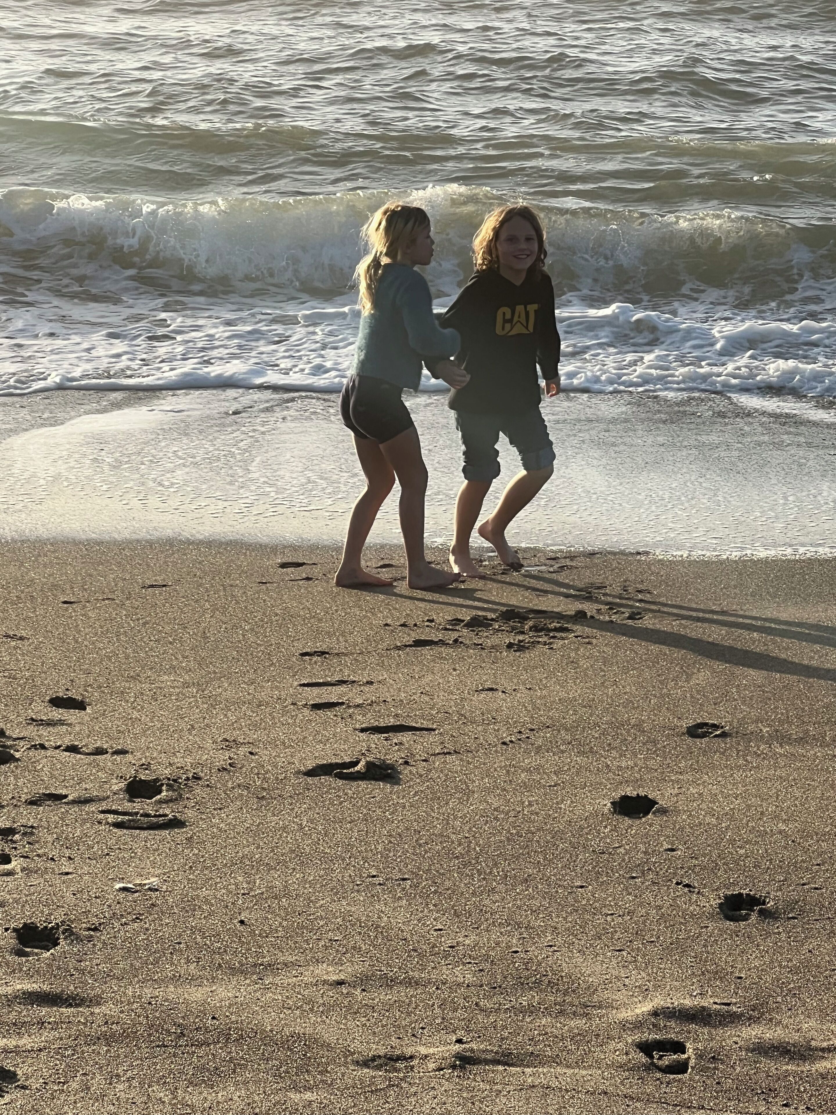 Two children walking on a sandy beach with footprints leading into the water.