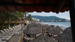 Blick auf einen Strand mit Reihen von Liegestühlen und Reetdachschirmen, einem Sandweg und einer Aussicht auf den Strand und entfernte Gebäude.