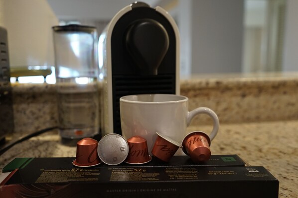 Una encimera de cocina con una cafetera, una taza blanca y una caja de cápsulas de café.