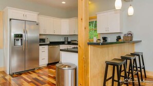 A modern kitchen with white cabinets, a stainless steel refrigerator, a microwave, and a wooden bar area with stools. - Newly Built Mountain Cabin With Views, Privacy And 10 Min. From Downtown! (Asheville)