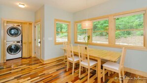 A room with a wooden floor, a dining table with chairs, a washing machine, and large windows with a view of greenery. - Newly Built Mountain Cabin With Views, Privacy And 10 Min. From Downtown! (Asheville)