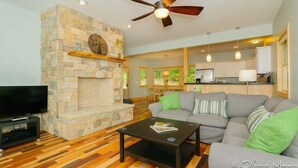 A living room with a stone fireplace, a ceiling fan, a television, and a sofa with green cushions. - Newly Built Mountain Cabin With Views, Privacy And 10 Min. From Downtown! (Asheville)