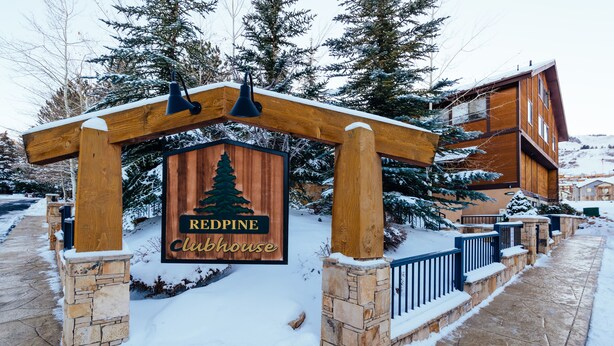 A wooden sign with a pine tree logo and the text "Redpine Clubhouse" in front of a building with stone pillars and a stone railing. - Deluxe Condo -complimentary shuttle to ski- Best location, comfort & style (Park City)