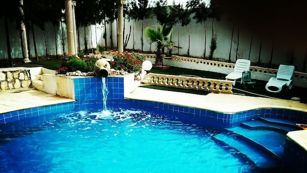 Una piscina con fuente, rodeada de un muro de piedra, con sillas de descanso y una palmera al fondo.