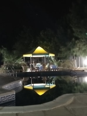 A gazebo with a glass enclosure over a pool, surrounded by lounge chairs and potted plants.