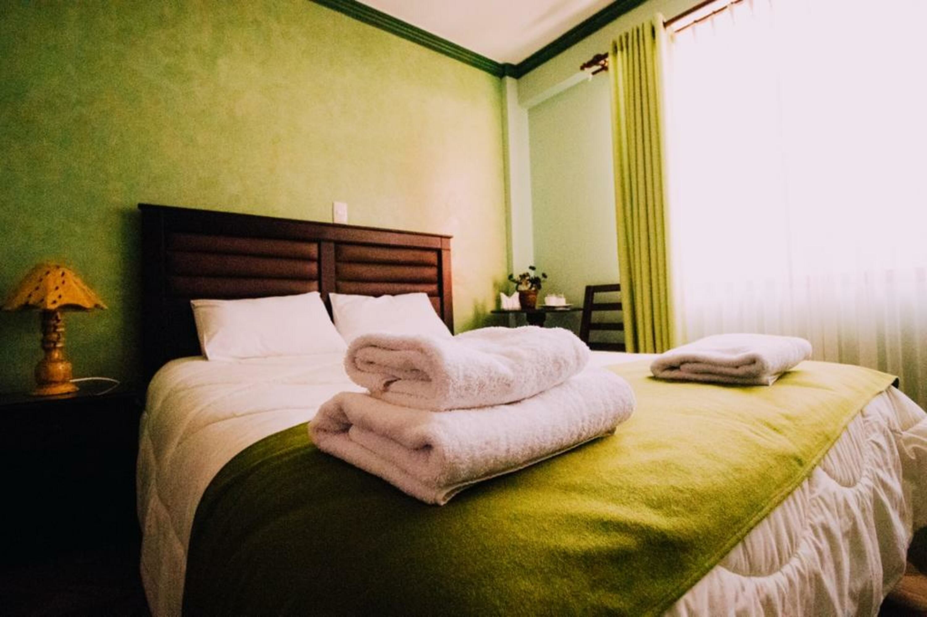 Una cama bien hecha con toallas dobladas, una pared verde y una lámpara en una mesita de noche.