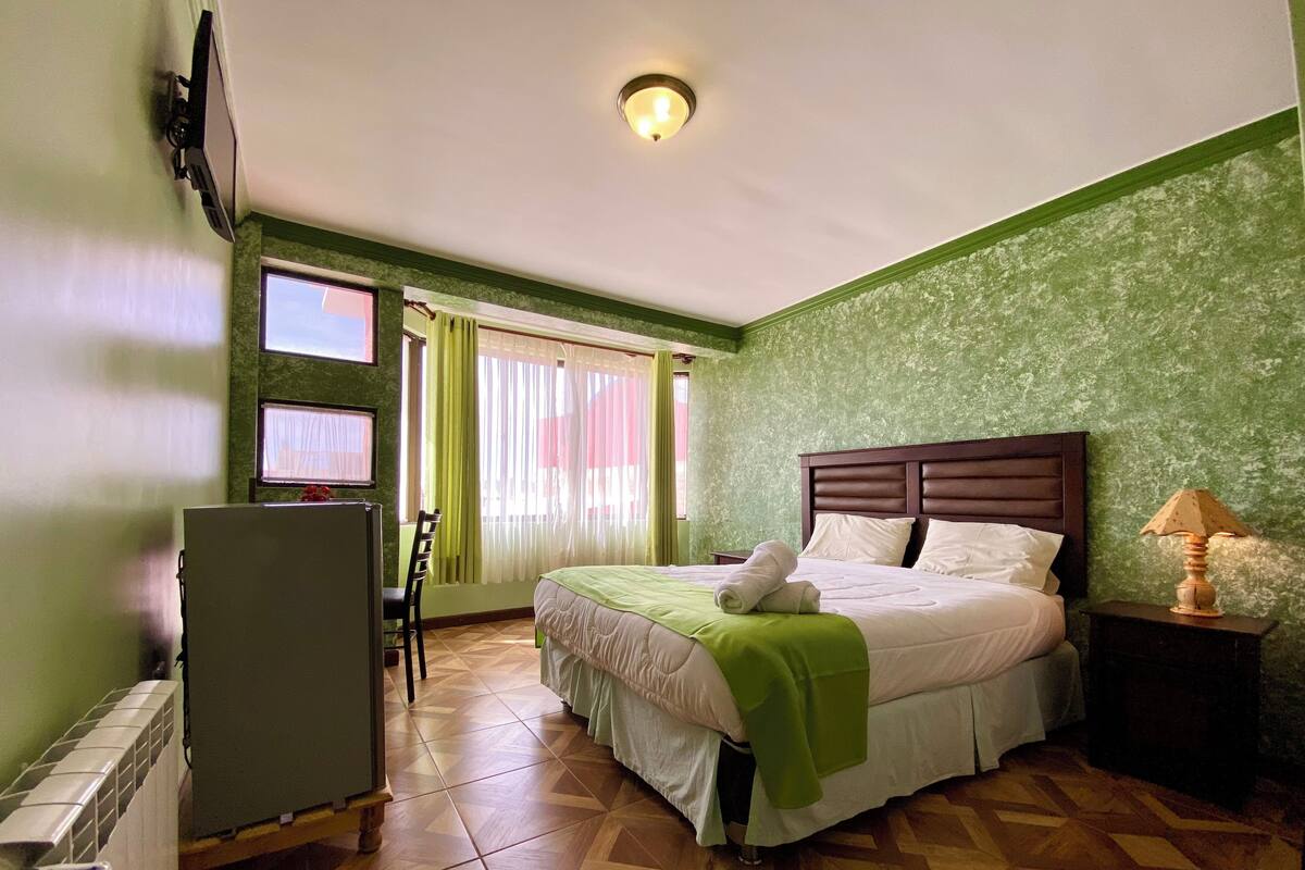 A hotel room with a bed, a desk, a chair, a lamp, and a television on the wall.