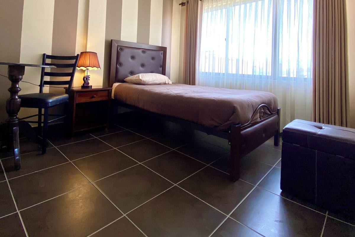 A hotel room with a bed, a chair, a table, a lamp, and a window with curtains.