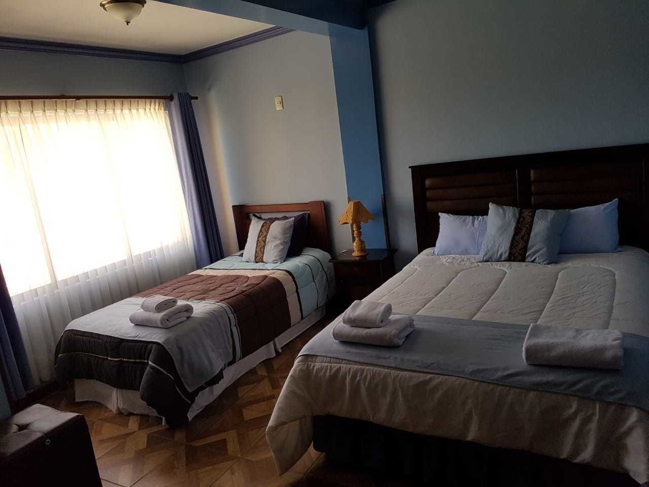 A hotel room with two beds, a nightstand, a lamp, and a window with curtains.