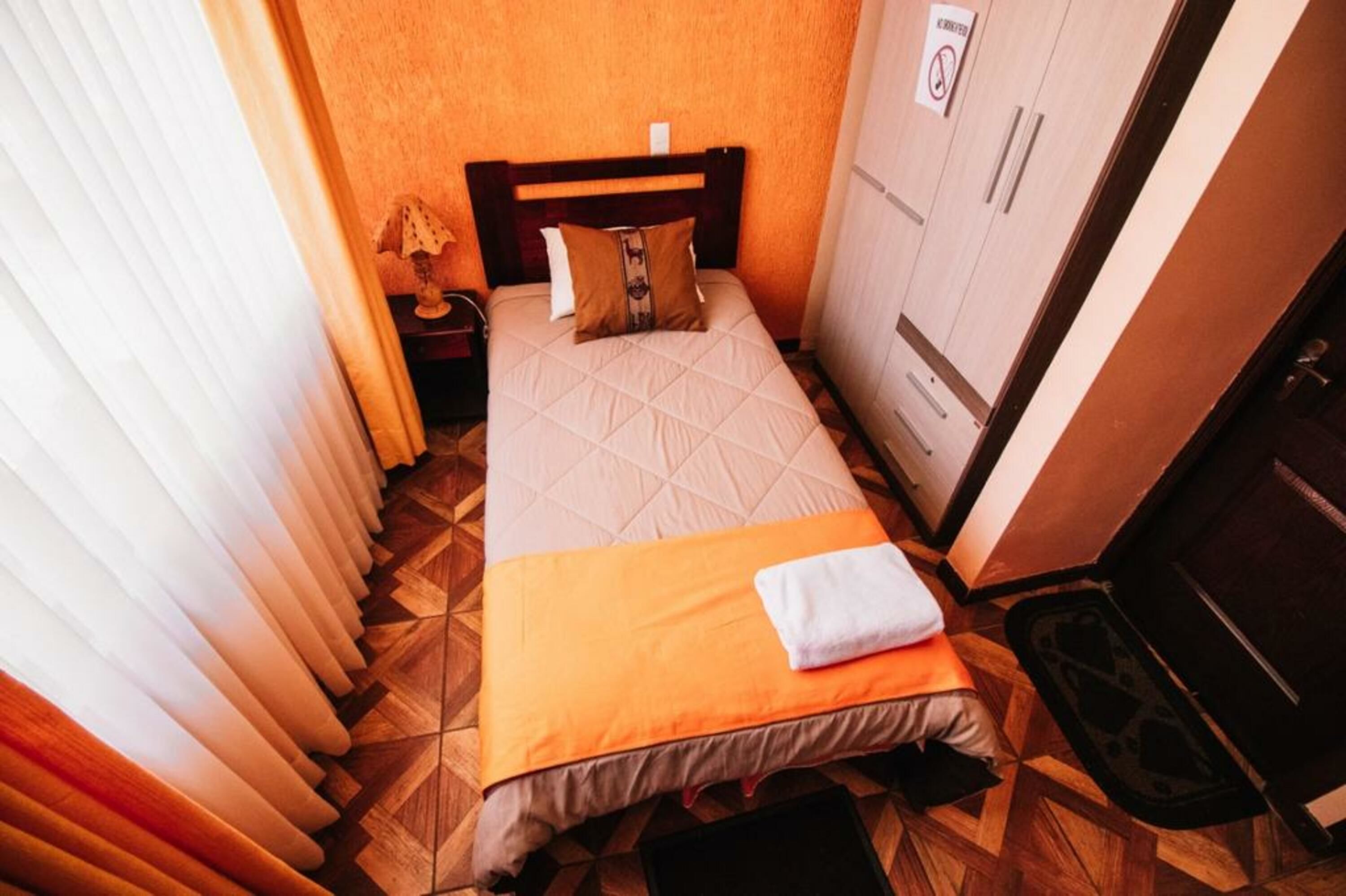 A small, neatly made bed with a quilted cover and a pillow, a bedside table with a lamp, and a wardrobe with a sign on it.
