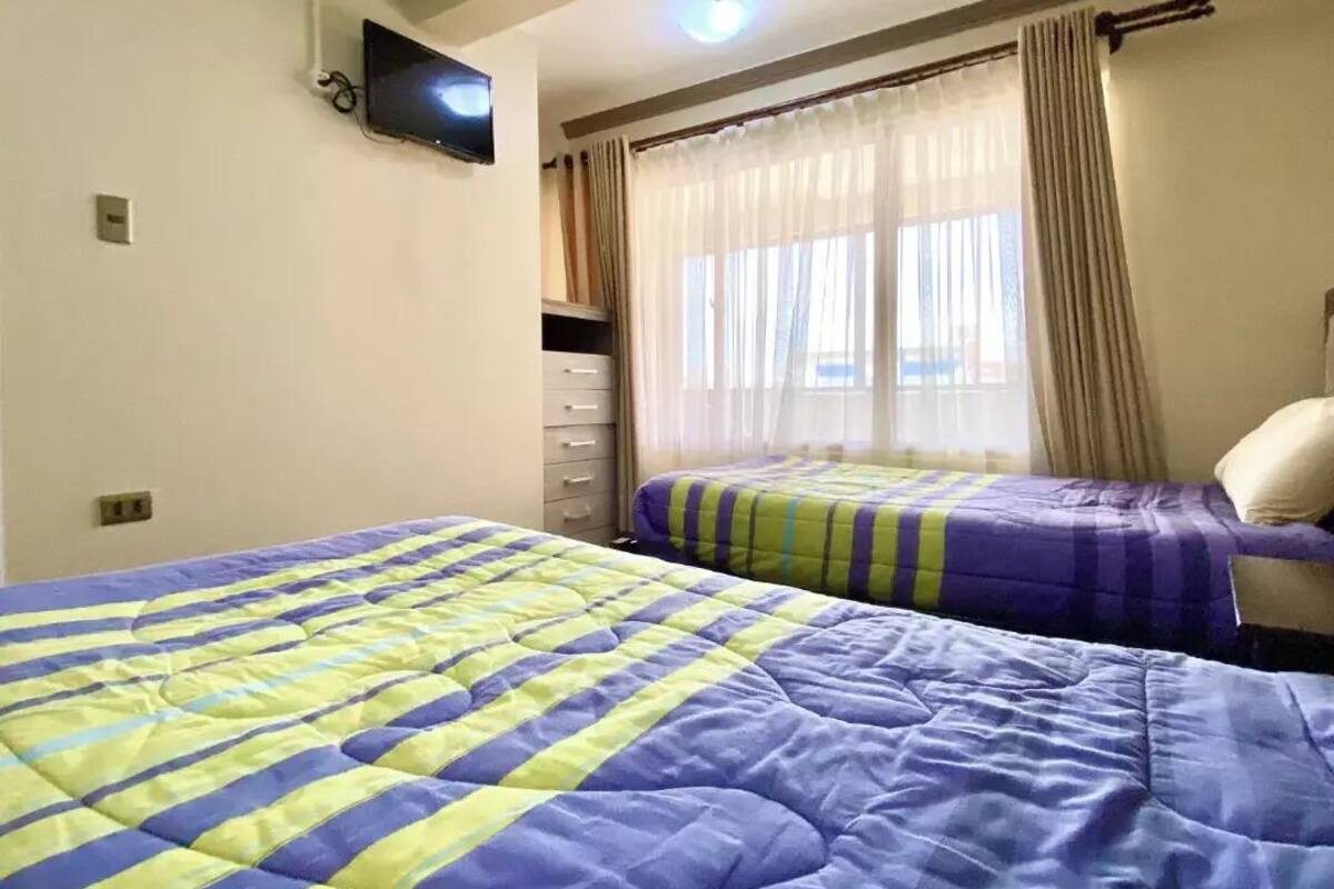 A hotel room with two beds, a television on the wall, a window with curtains, and a ceiling light.