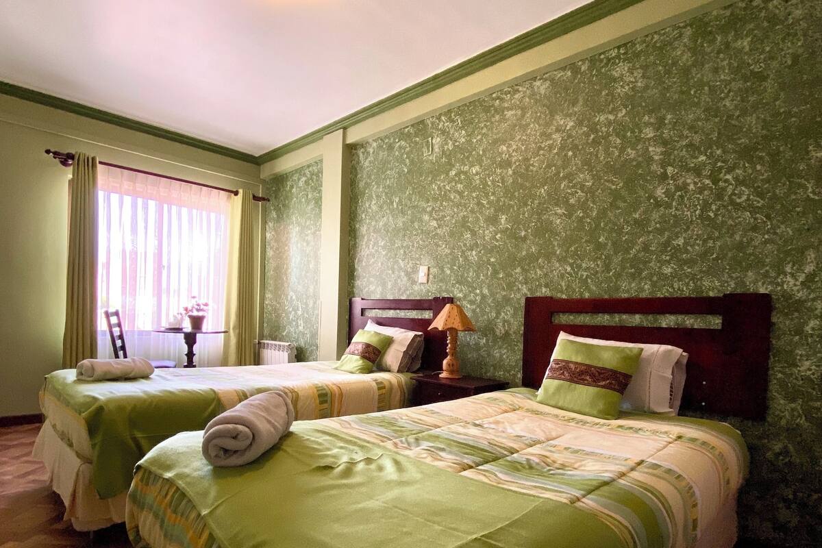 A hotel room with two beds, a window with curtains, a wooden floor, and a green textured wall.