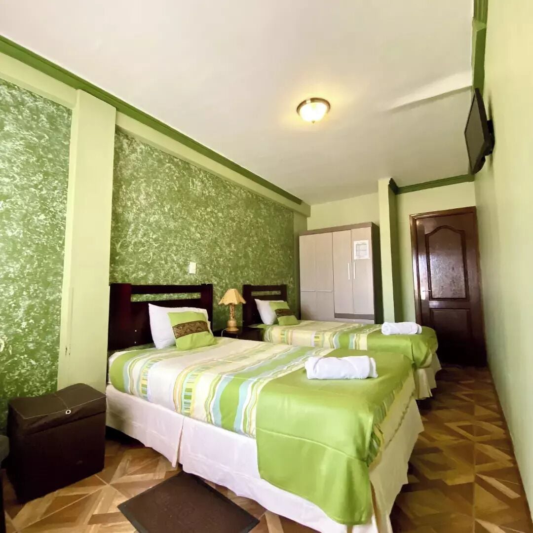 Habitación de hotel con dos camas, paredes de color verde con estampado, suelo de madera, un armario y un televisor en la pared.