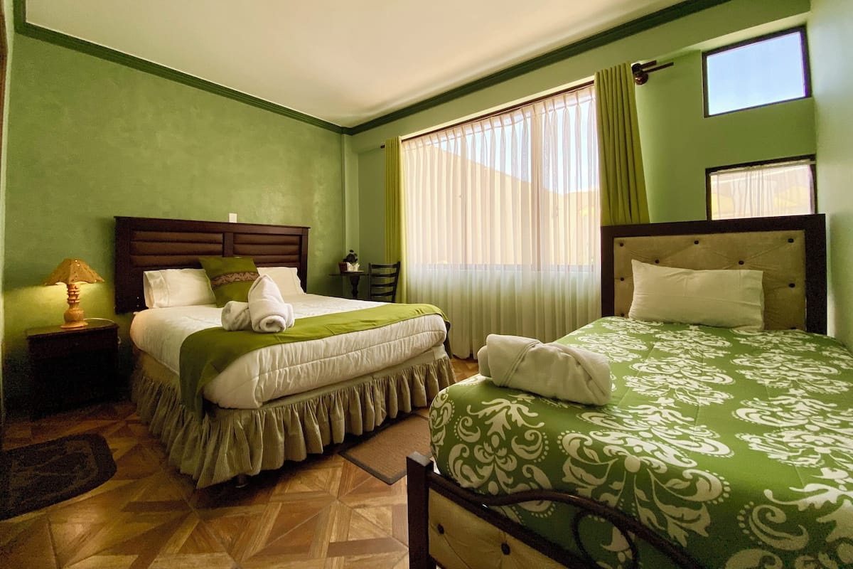 A hotel room with two beds, a wooden headboard, a nightstand with a lamp, a small table with a plant, and a window with curtains.