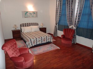 A bedroom with a bed, two nightstands, a red armchair, a matching ottoman, a wooden floor, and a window with curtains. - Casa do Abade - Solar de Goujoim (Armamar)