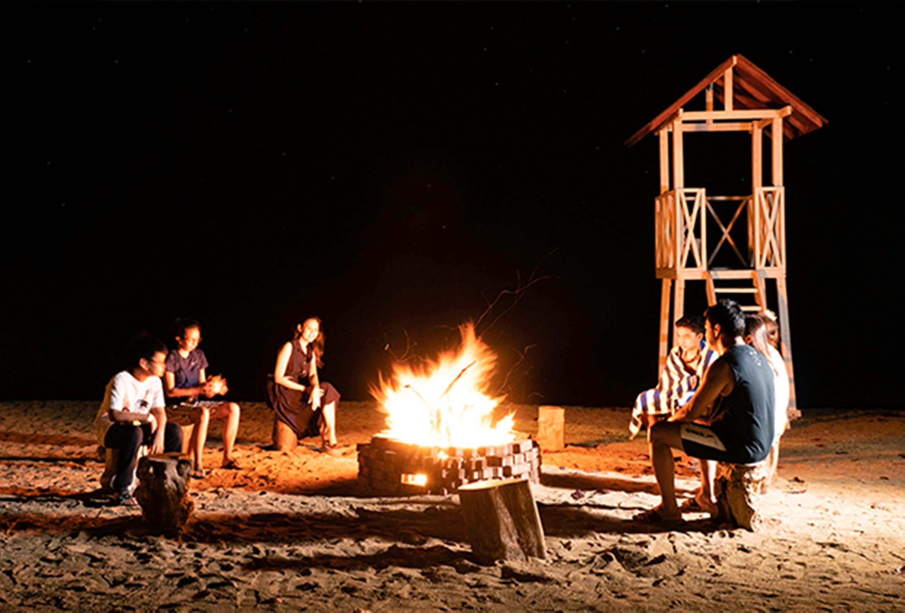 A group of people gathered around a campfire on a sandy beach at night, with a wooden structure in the background.