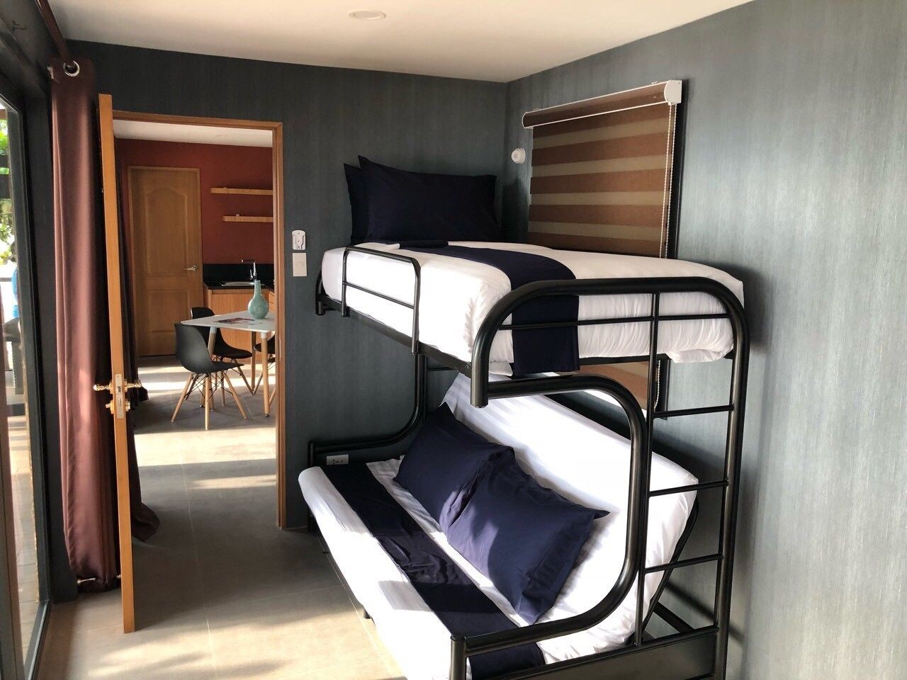 Une chambre avec un lit superposé, un bureau et une chaise, une porte donnant sur un coin repas et une fenêtre avec des rideaux.