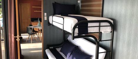 Habitación con literas, un escritorio y una silla, una puerta que da a un comedor y una ventana con cortinas.