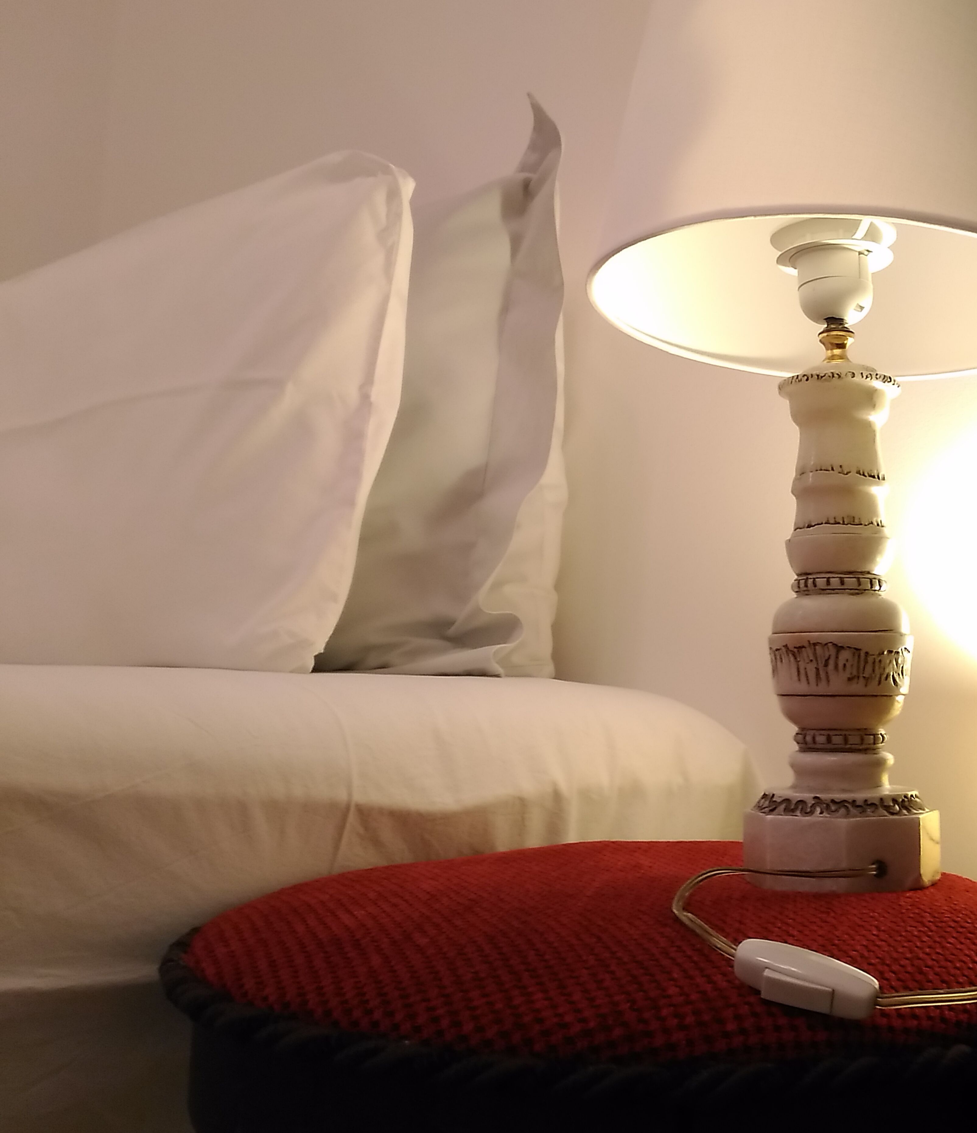 Ein Bett mit weißen Leinen, einem roten Kopfkissen und einem Nachttisch mit Lampe und Netzteil.
