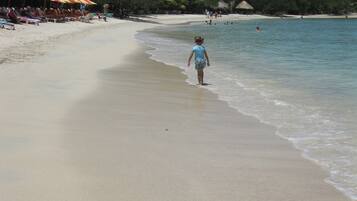 Una playa con un niño caminando hacia el agua, sombrillas y gente disfrutando en el fondo.