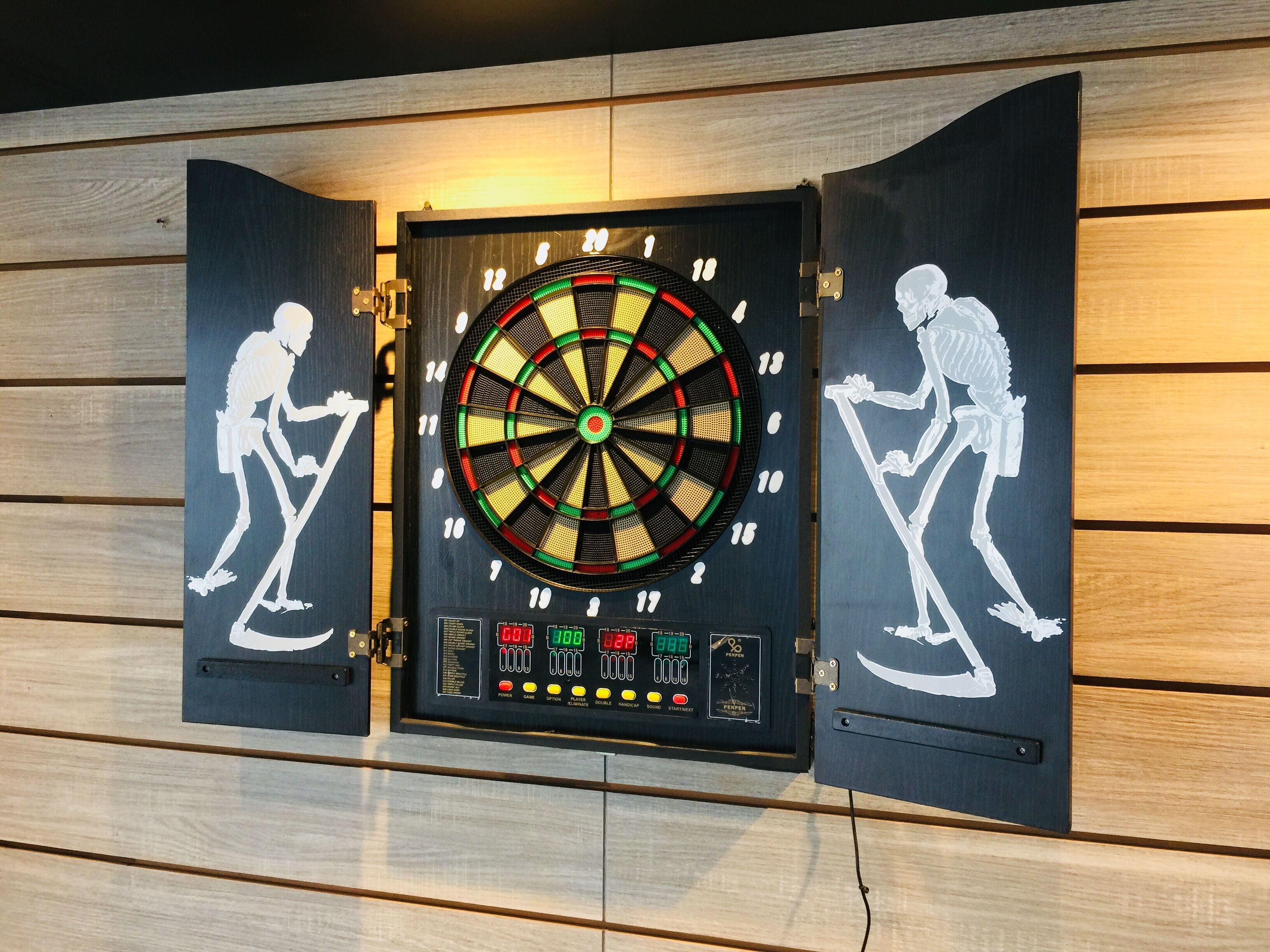 Un tablero de dardos con segmentos iluminados y una pantalla digital de puntuación, montado sobre una pared de madera.