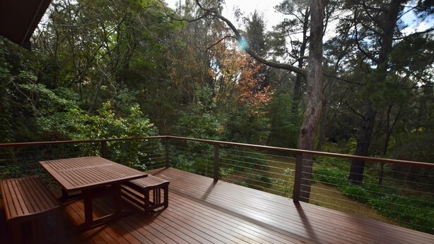 Una terraza de madera con una mesa y bancos, rodeada de árboles y una barandilla.