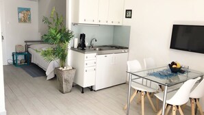 Cocina compacta con armarios blancos, un lavamanos y una zona de comedor con mesa de vidrio y sillas blancas.
