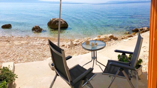 Un patio con dos sillas y una mesa, con vista a una playa de aguas azules y rocas.