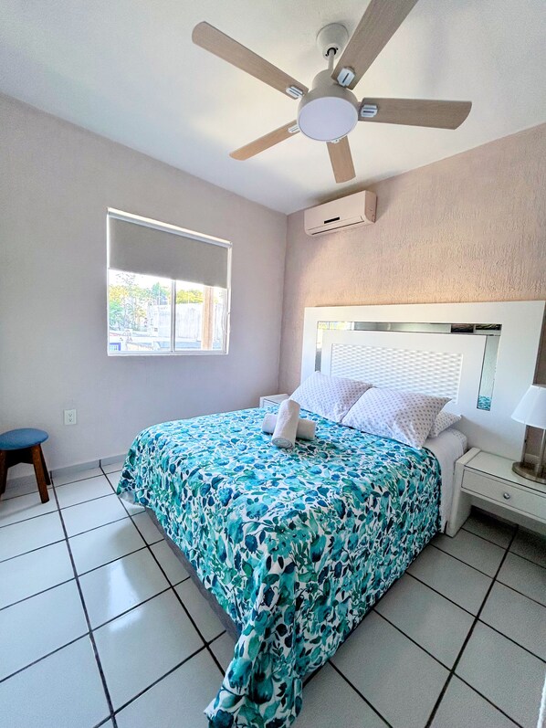 A bedroom with a bed, a ceiling fan, an air conditioning unit, a bedside table with a lamp, and a window with blinds.