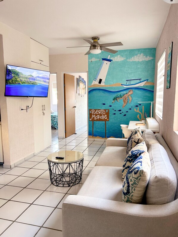 A living room with a television, a ceiling fan, a mural of a lighthouse, and a sign reading 'Puerto Morelos'.