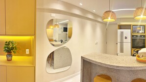 A modern kitchen with a curved granite countertop, a large mirror, and a hanging pendant light. - Vacanze - edf. Port Ville III apt 309, rooftop pool with panoramic views. (Maceió)