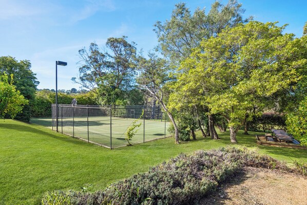 A tennis court with a chain-link fence, surrounded by well-maintained grass and trees. - Tecnevin of Shoreham, Mornington Peninsula (Shoreham)