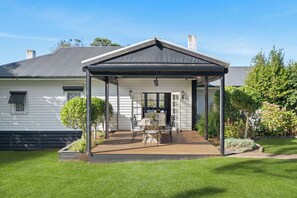 Une terrasse couverte avec une table et des chaises, un petit jardin et une maison blanche avec un toit gris.