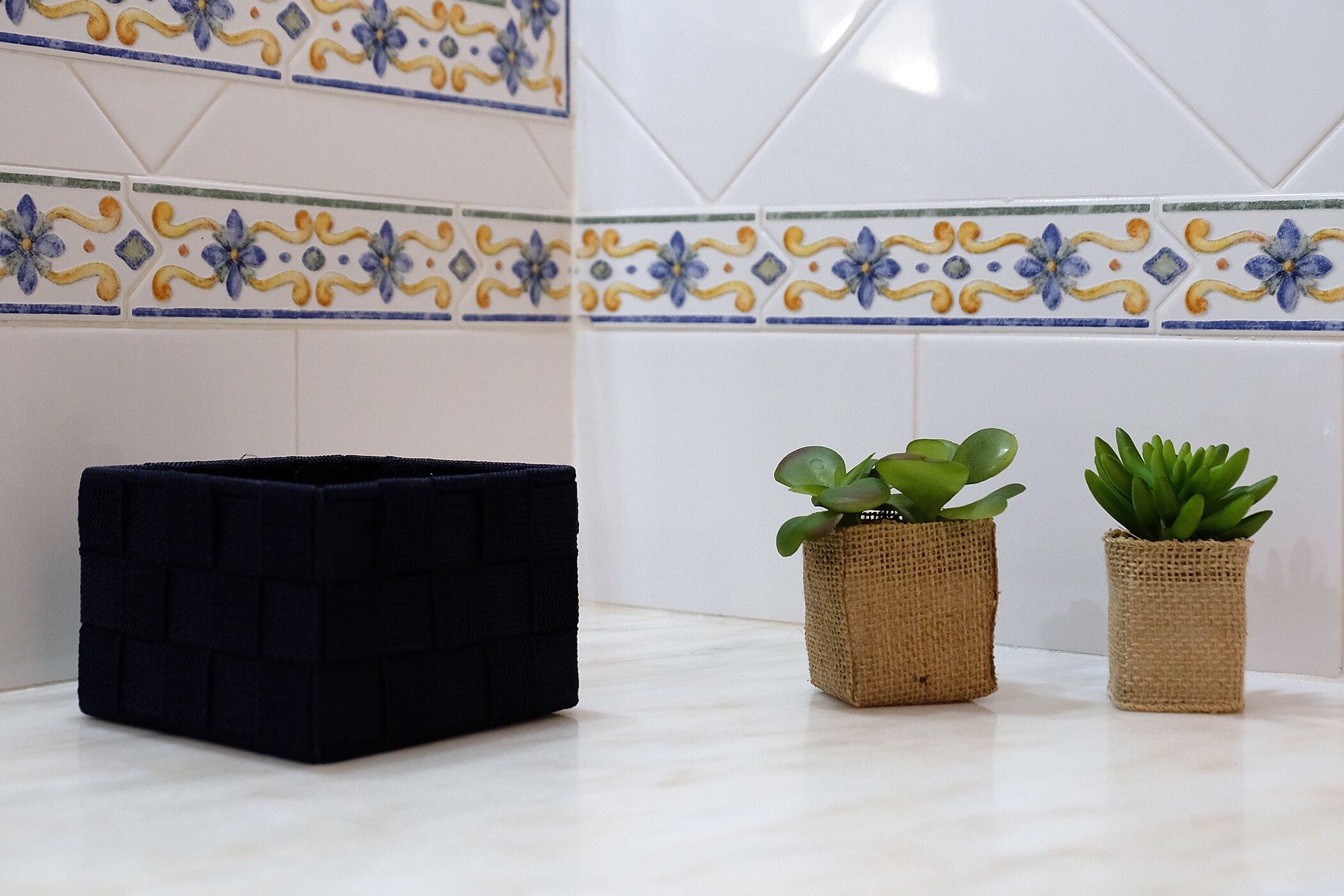 Een kamer met een betegelde spatwand met een bloemmotief, een zwarte opbergdoos en twee kleine potplanten.