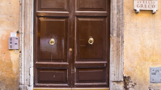 A large, ornate double door with a brass doorknob, set in a weathered yellow wall with a plaque above it.