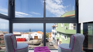 A room with two grey upholstered chairs and a purple ottoman, a large window with a view of buildings and the sea. - Luxury villa with panoramic ocean view (Guía de Isora)