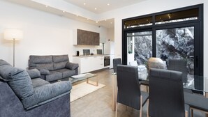 A modern living room with a grey sofa, a glass coffee table, and a dining area with chairs. There is a kitchen in the background with white cabinets and a large window showing a rocky landscape. - Luxury villa with panoramic ocean view (Guía de Isora)