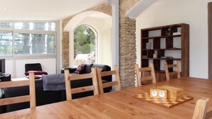 Un coin repas avec une table en bois, des chaises et un jeu d’échecs. On aperçoit une bibliothèque et un canapé à l’arrière-plan.