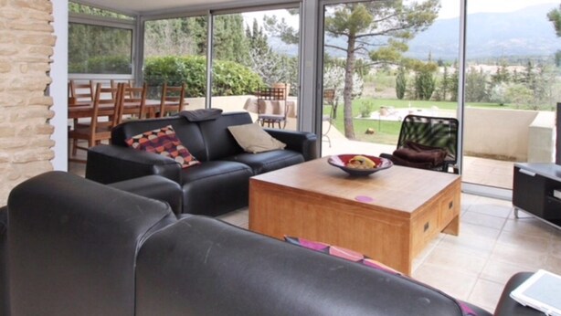 Un salon moderne avec un canapé en cuir noir, une table basse en bois et un coin repas comprenant une table et des chaises. De grandes fenêtres offrent une vue sur l’extérieur.