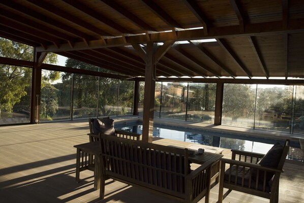 A covered outdoor area with a pool, wooden furniture, and large windows offering a view of trees and a landscape. - Casa de Campo el Encinar - Paddleball, Pickleball, Swimming Pool and Barbecue (Santa Cruz del Retamar)