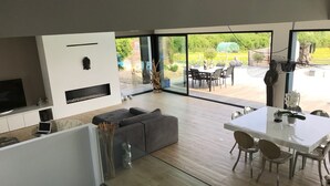 A modern living room with a sofa, a dining table with chairs, a TV, and a large window overlooking a garden. - Architect Villa with heated pool  (Séné)