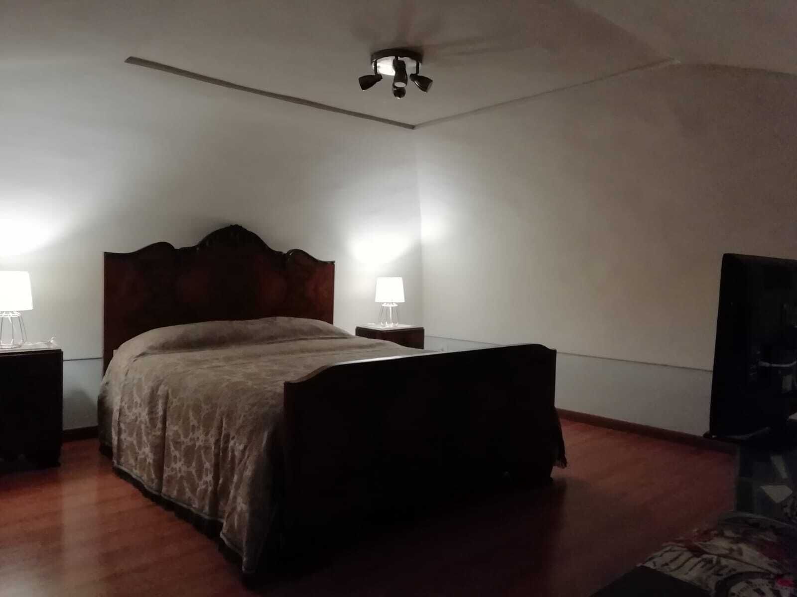 Une chambre à coucher avec un lit en bois, deux tables de chevet avec des lampes, un ventilateur de plafond et un téléviseur fixé au mur.