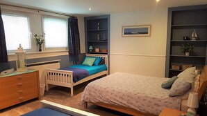 Une chambre avec un lit simple, une commode, une table de chevet, une fenêtre avec des rideaux, un radiateur et des étagères intégrées décorées de divers objets.